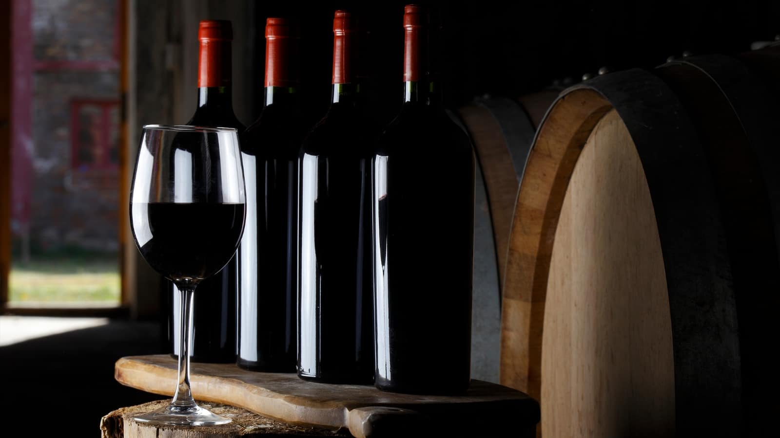 a glass of red wine stands against the background of bottles of wine and wine barrels