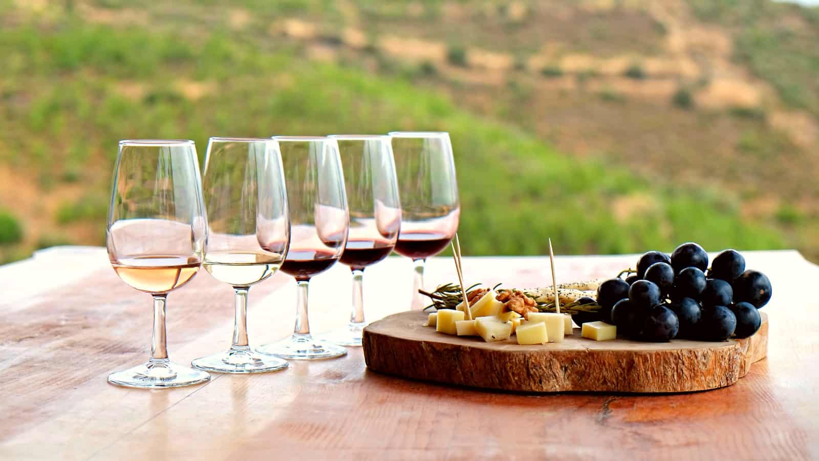 Close-up of five glasses with different types of red and white wine, a wooden plate full of cheese and grapes on a wooden table with a picturesque background