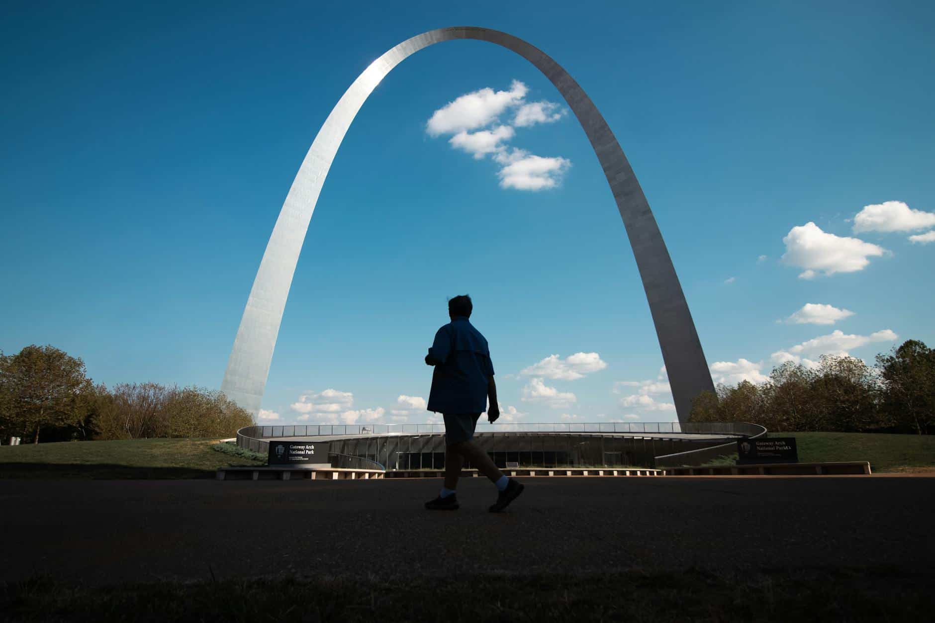 Silhouette of a person walking by the iconic Gateway Arch under a clear blue sky.