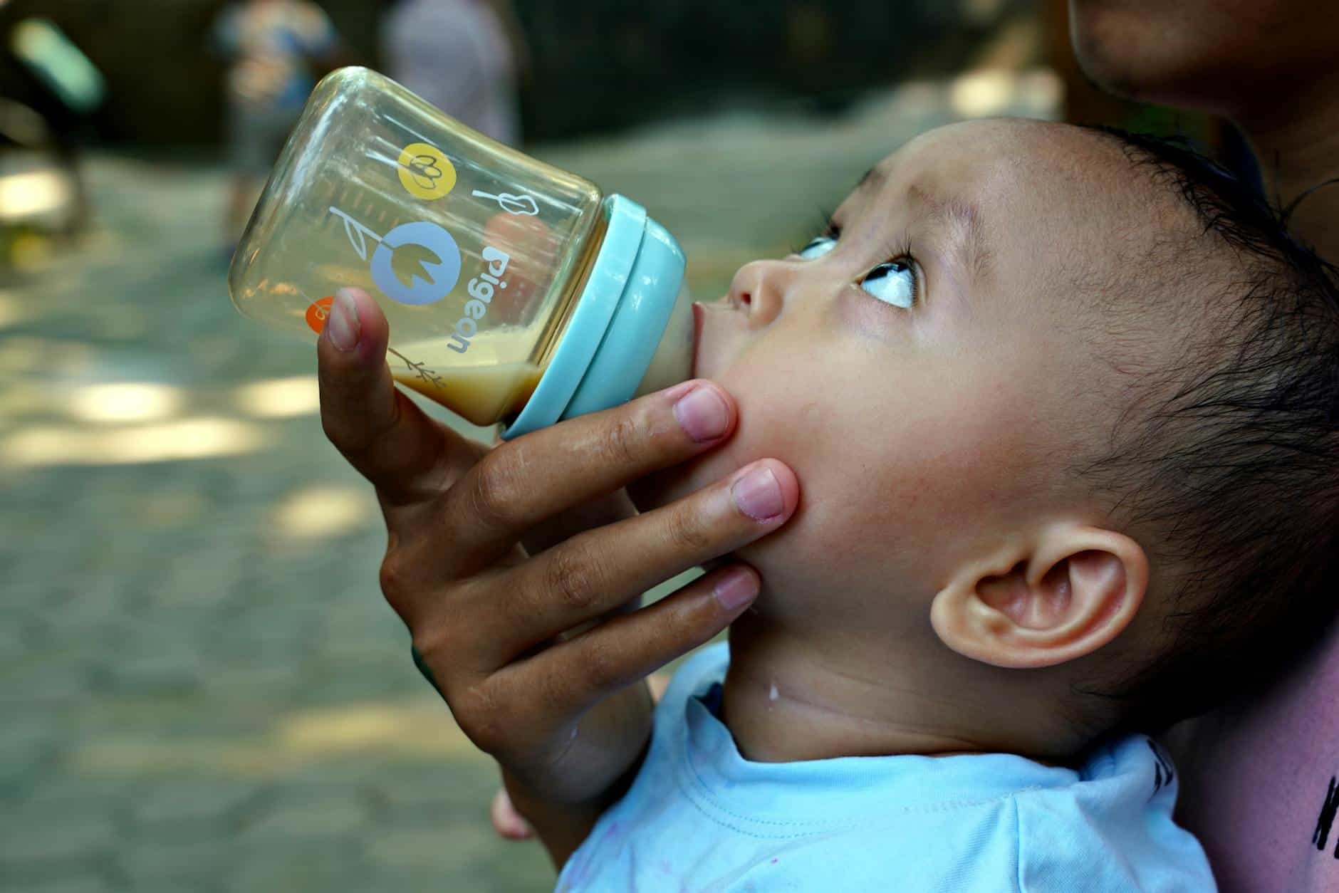 15 Road Trip Car Organizers for Traveling With Kids 14 A baby drinking from a bottle outdoors, enjoying a sunny day with a caregiver.