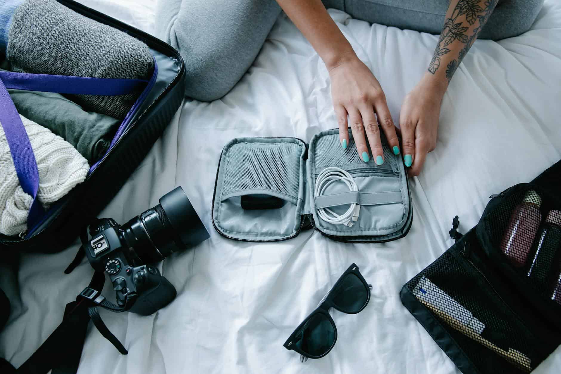 15 Road Trip Car Organizers for Traveling With Kids 6 Woman with tattoos packs electronics, camera, and sunglasses in a suitcase on a bed.