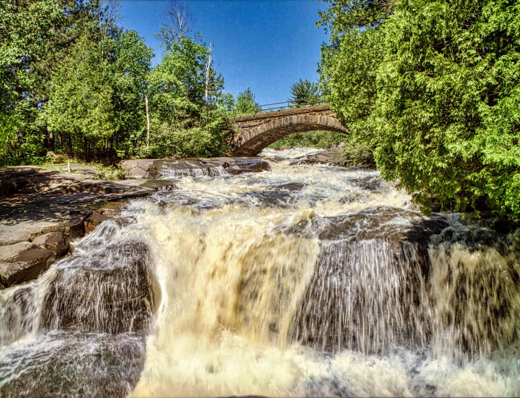 A picturesque waterfall flowing under a historic bridge in Duluth, MN, surrounded by lush greenery.