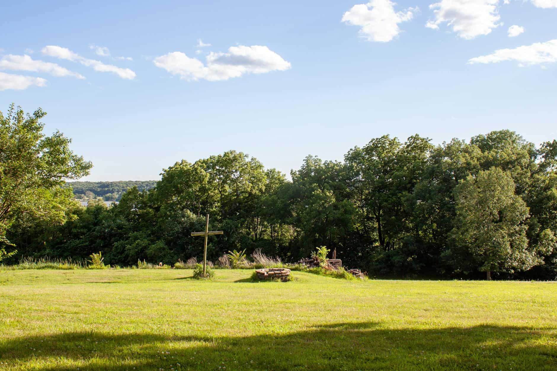 A serene countryside scene in Illinois featuring a lush green field, vibrant trees, a cross, and a clear blue sky.