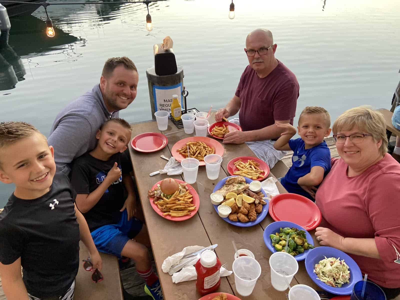 People enjoy a meal at a lakeside picnic table, with food and drinks on the table, and the water in the background.