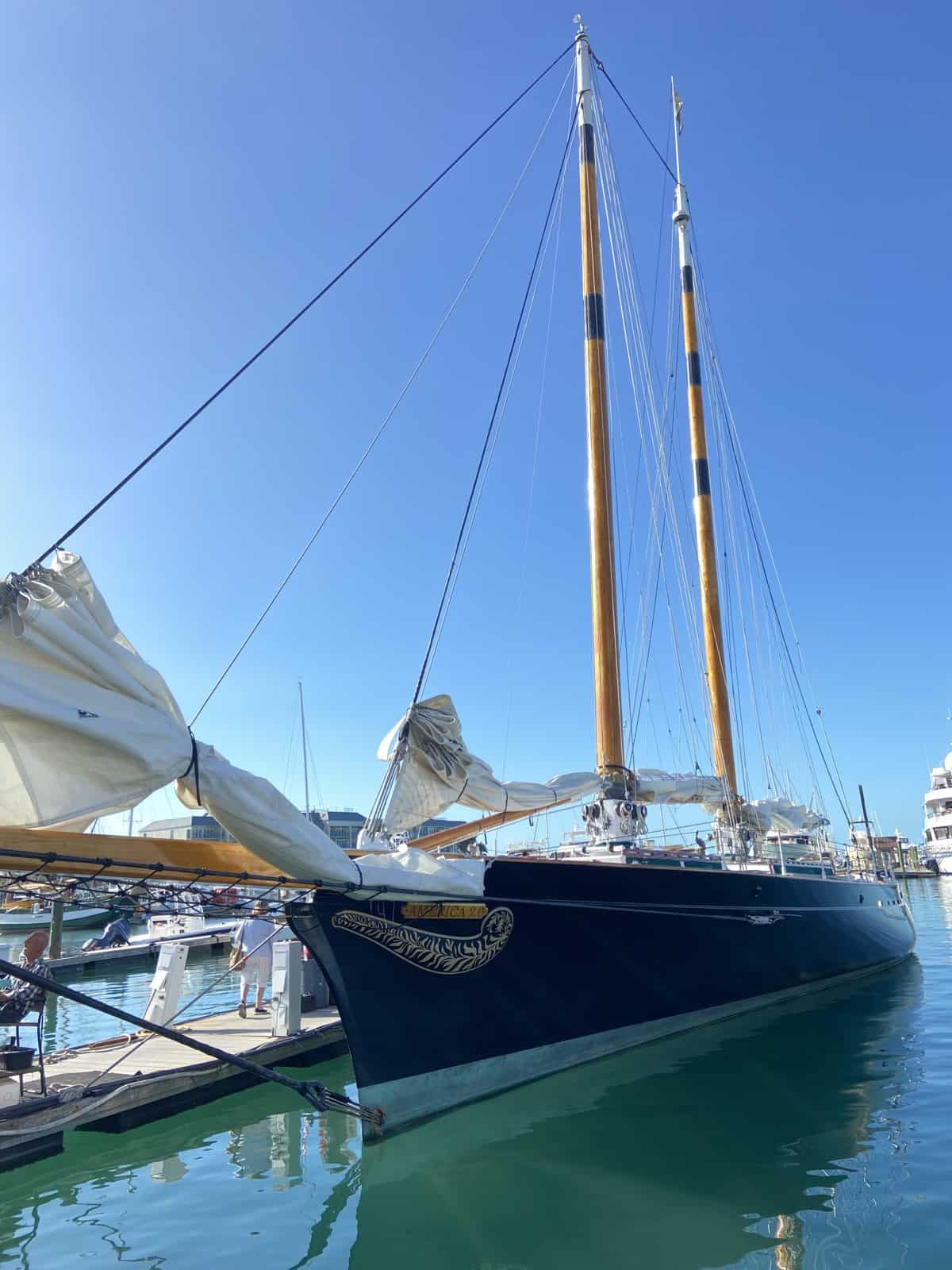 A large, dark blue sailboat with two tall masts is docked in a marina on a clear, sunny day. The boat is the main subject, with calm water and other boats in the background.
