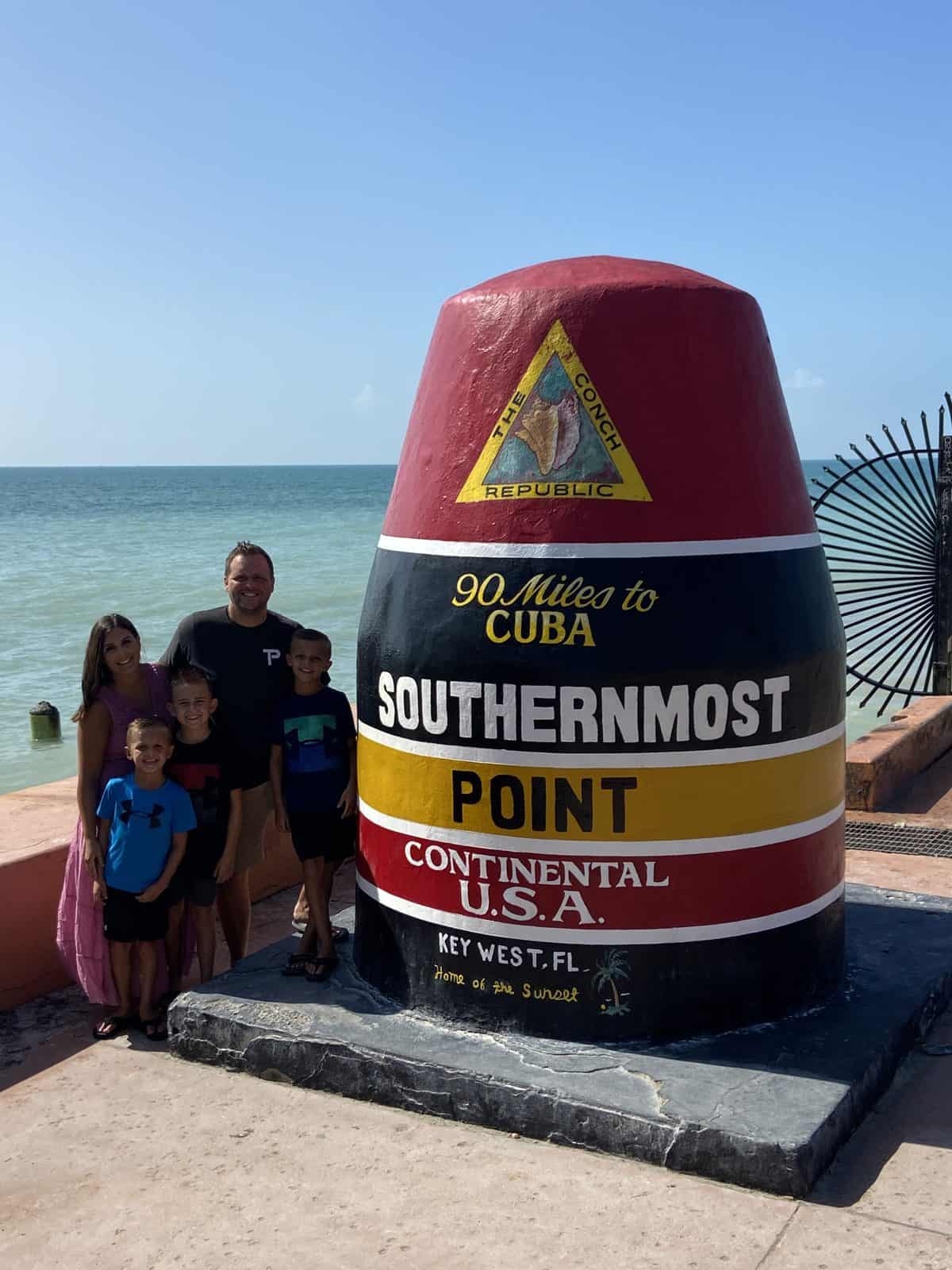 People stand beside a large, colorful monument marking Southernmost Point in Florida, with the ocean in the background.