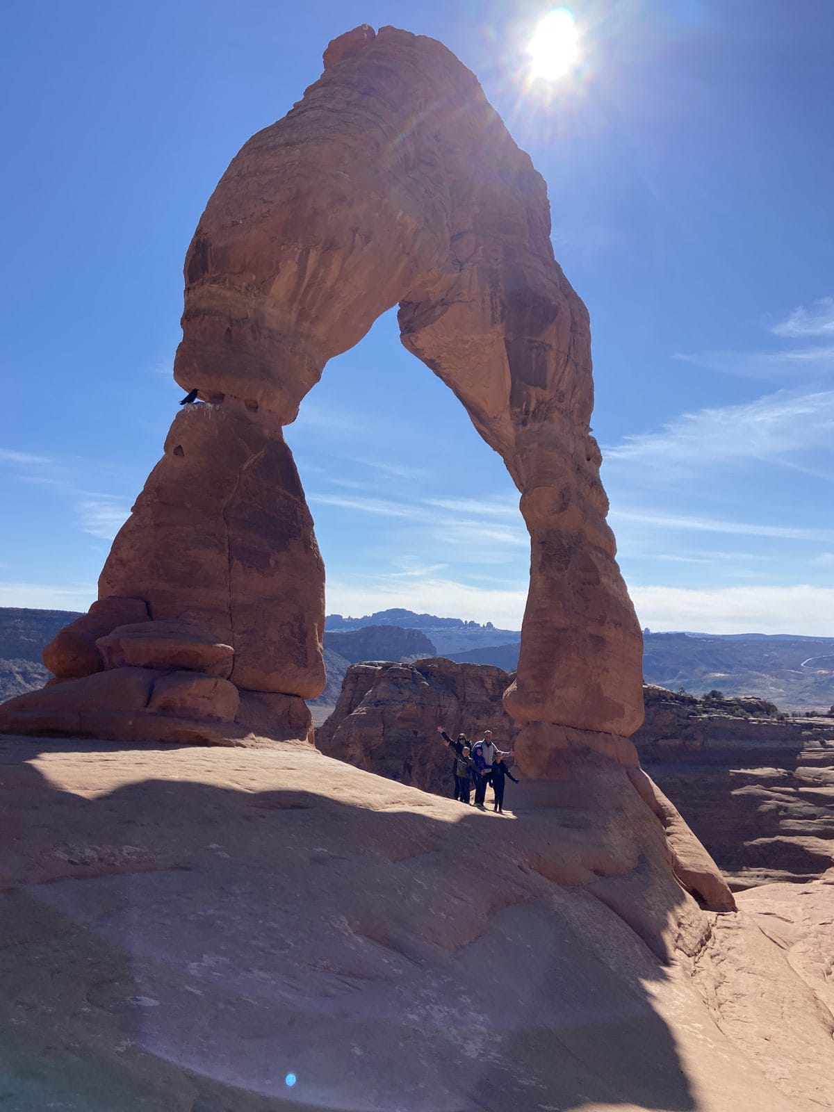 A large, red sandstone arch under a bright sun, with two people standing at its base in a desert landscape.