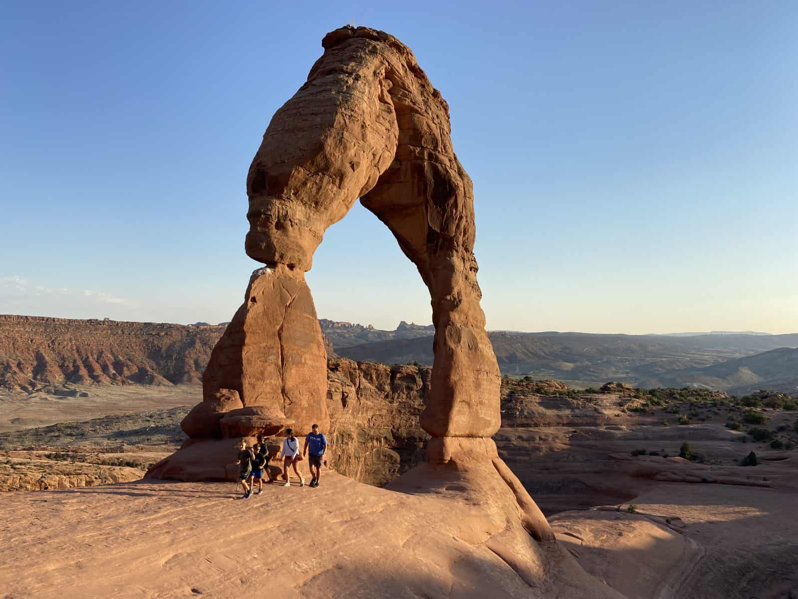 A group of people stands on a rock formation, with a large arch-shaped rock formation in the background under a clear sky.