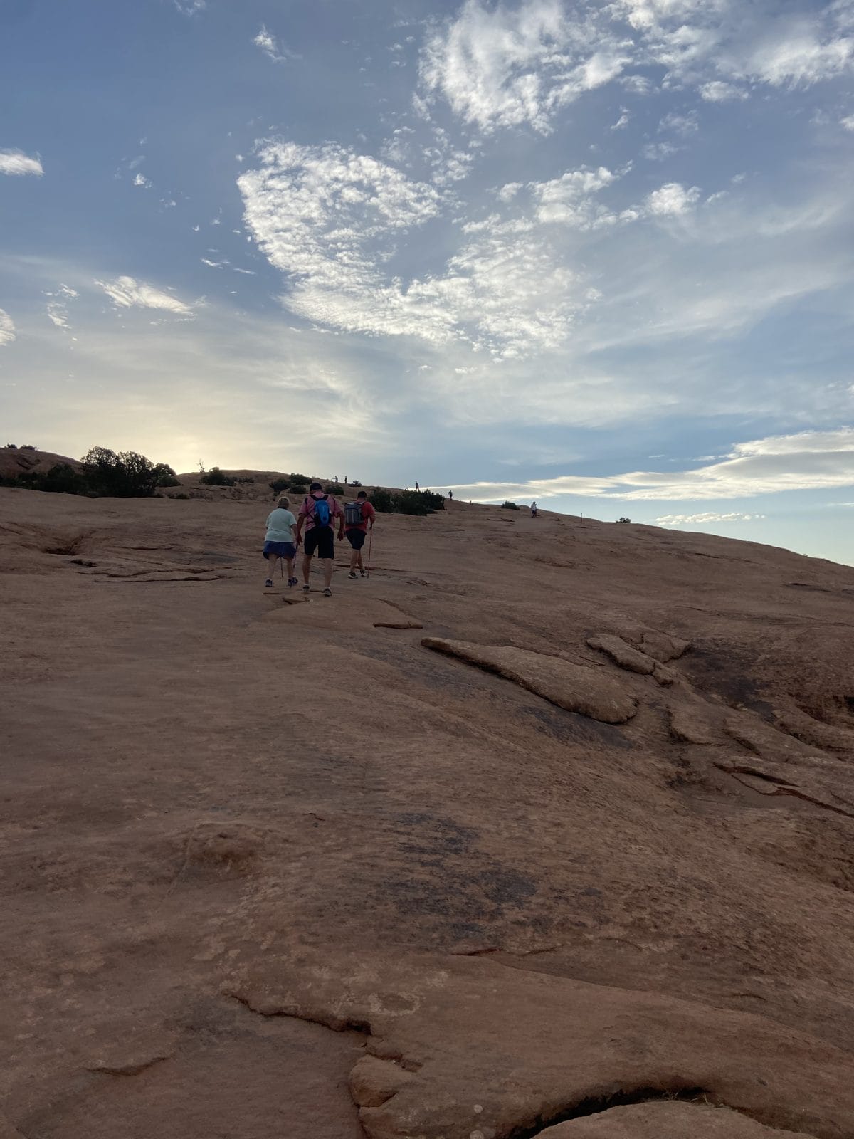 People hike up a large, rocky hill under a partly cloudy sky.