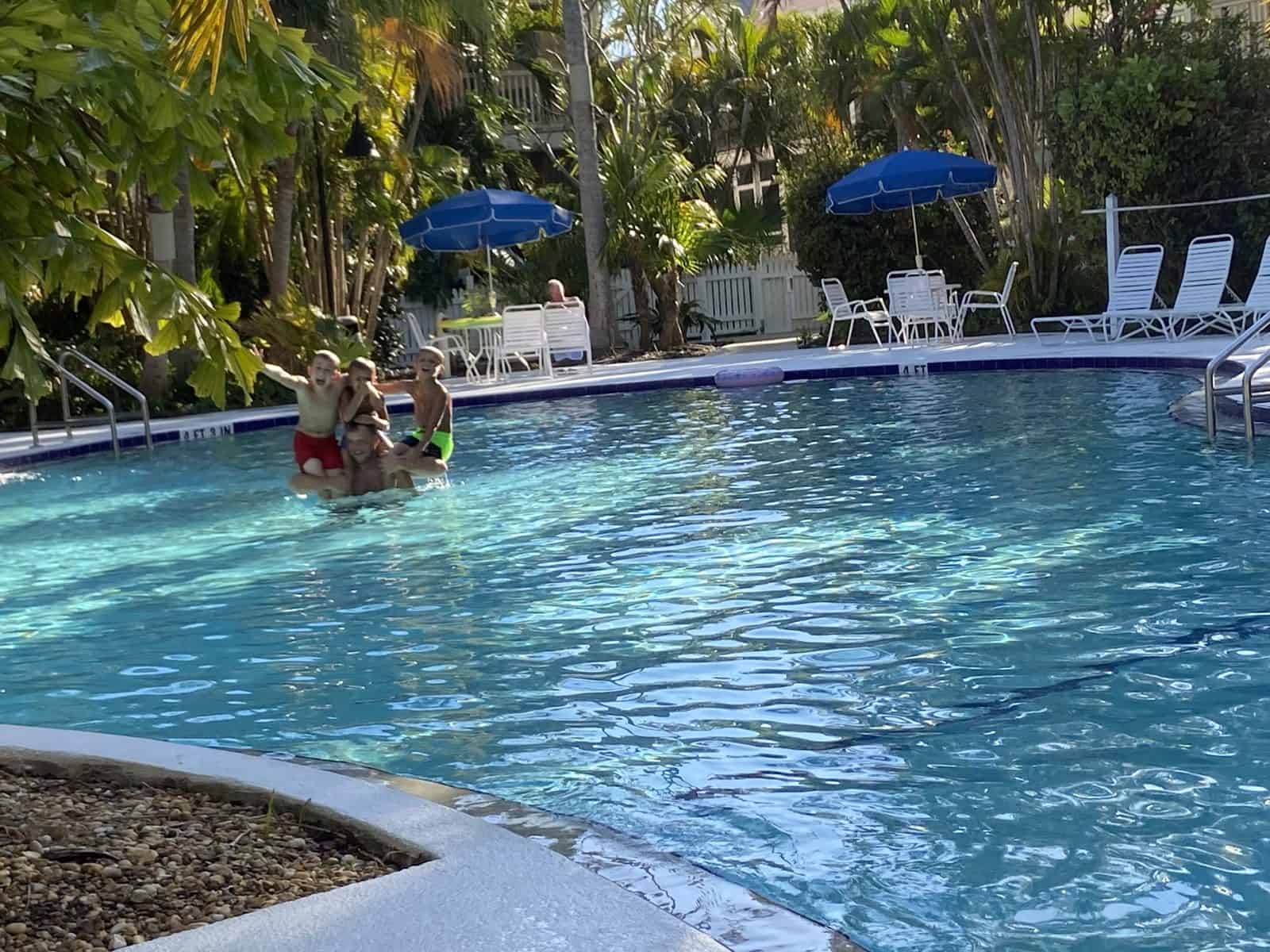 People in a pool, surrounded by palm trees and lounge chairs under blue umbrellas.