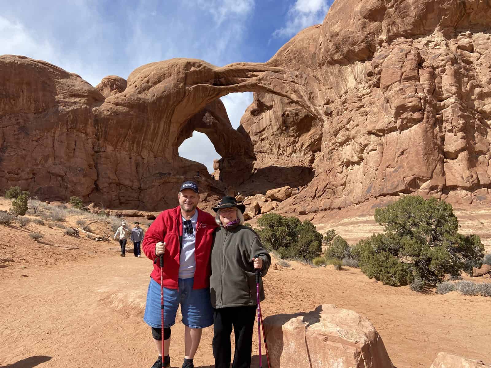 Two people pose in front of a large rock arch in a desert landscape, with a clear sky and other hikers in the background.
