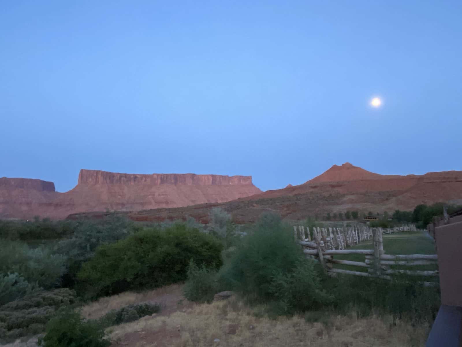 A dusk landscape with a moon, red rock formations, and a wooden fence in the foreground.