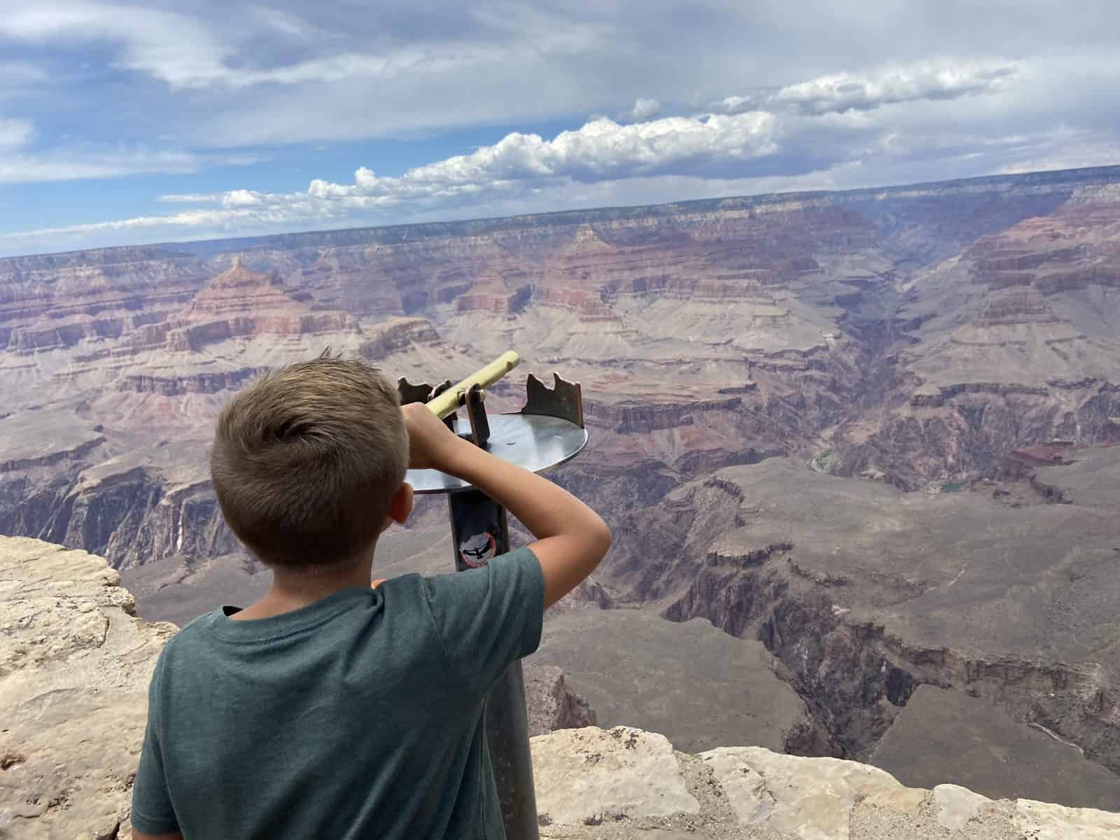 A boy stands on a cliff overlooking a vast, layered canyon, using a telescope to view the expansive landscape.