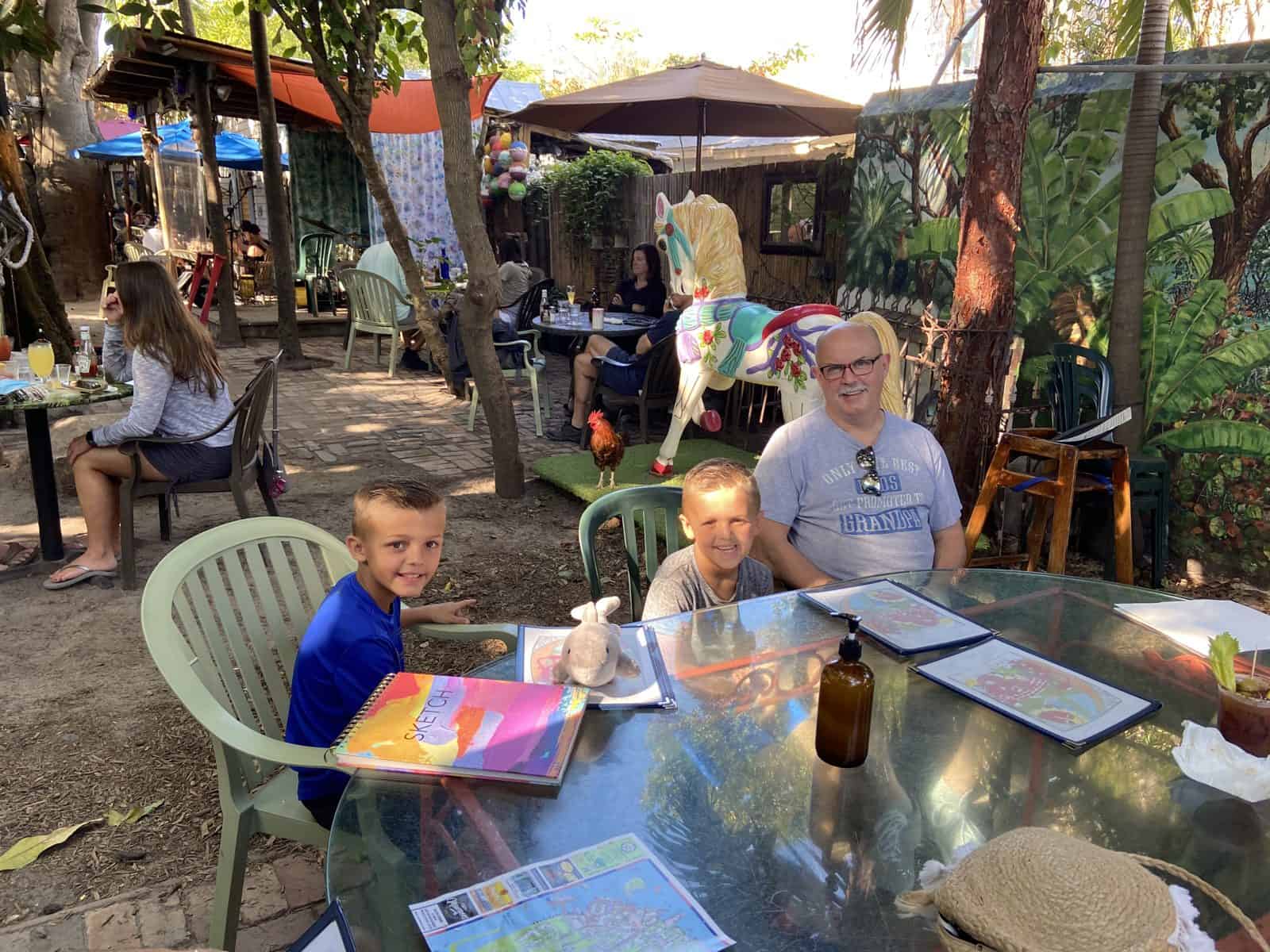 People at an outdoor cafe, seated at a table with menus and drinks, surrounded by trees and other patrons.