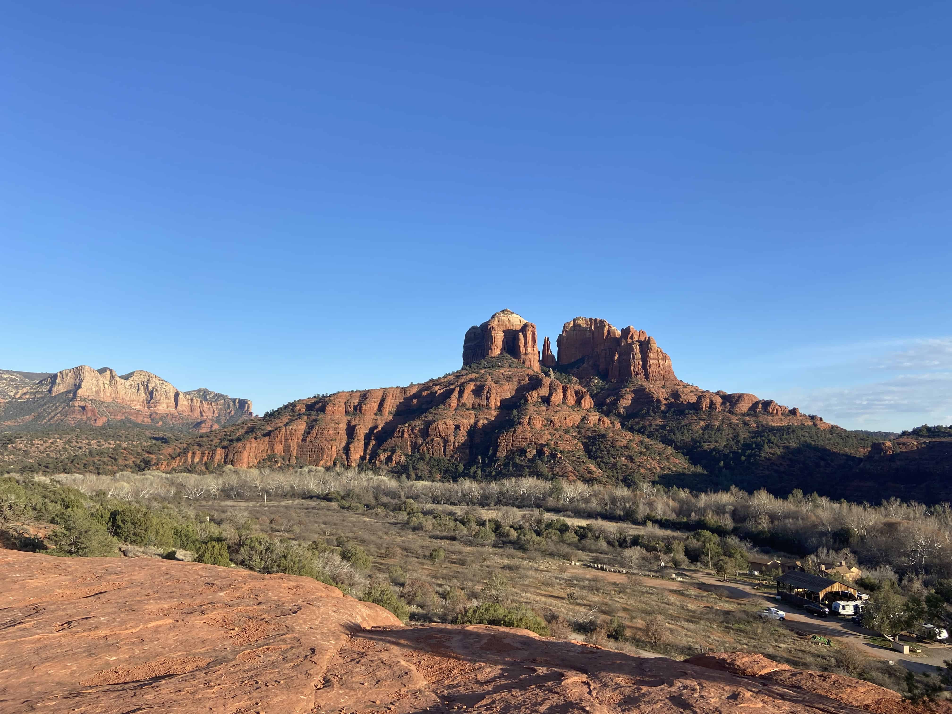 A scenic red rock landscape under a clear blue sky, with a distant mountain range and a small settlement in the valley.