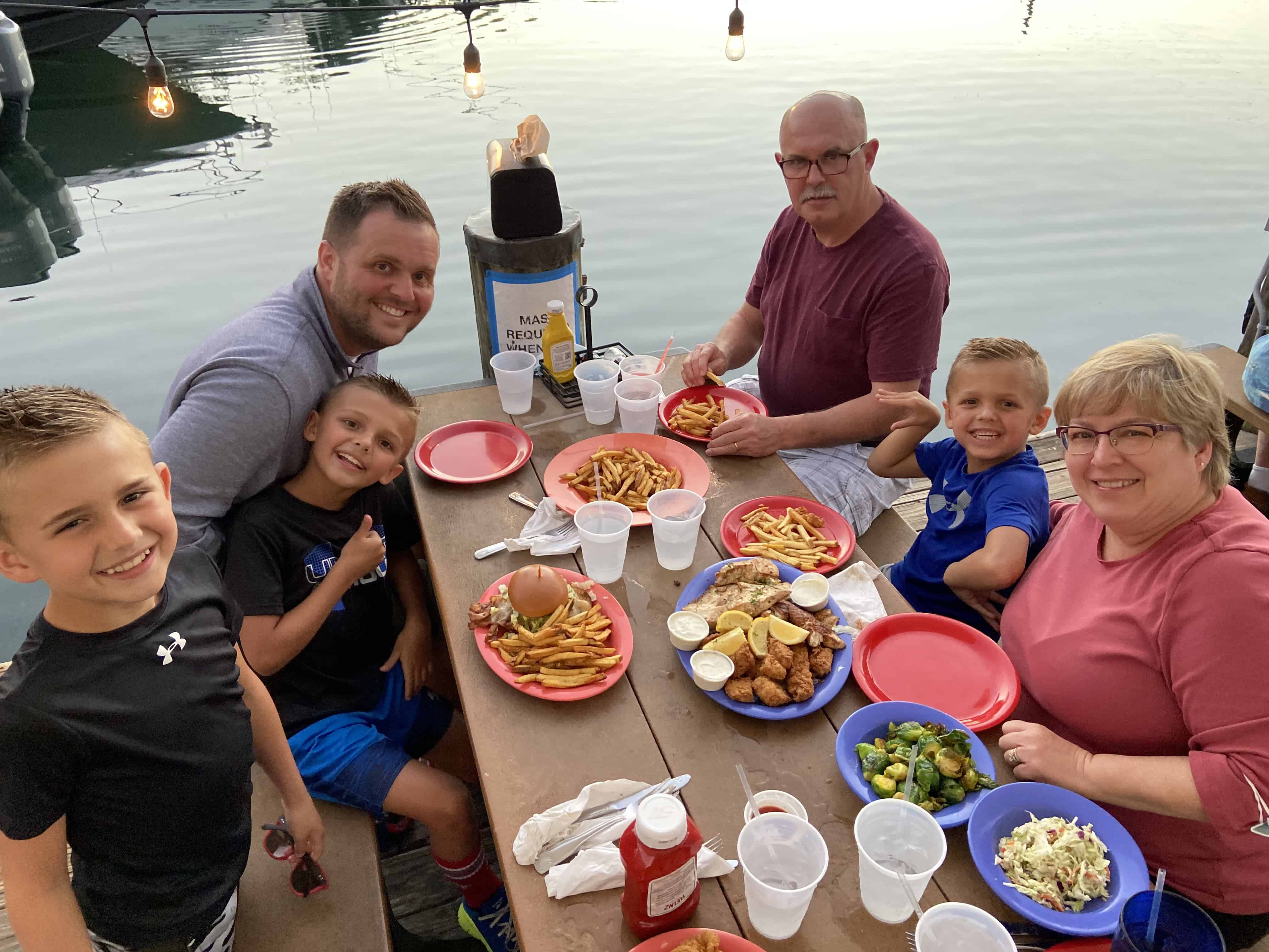 People enjoy a meal at a lakeside picnic table, with food and drinks on the table, and the water in the background.