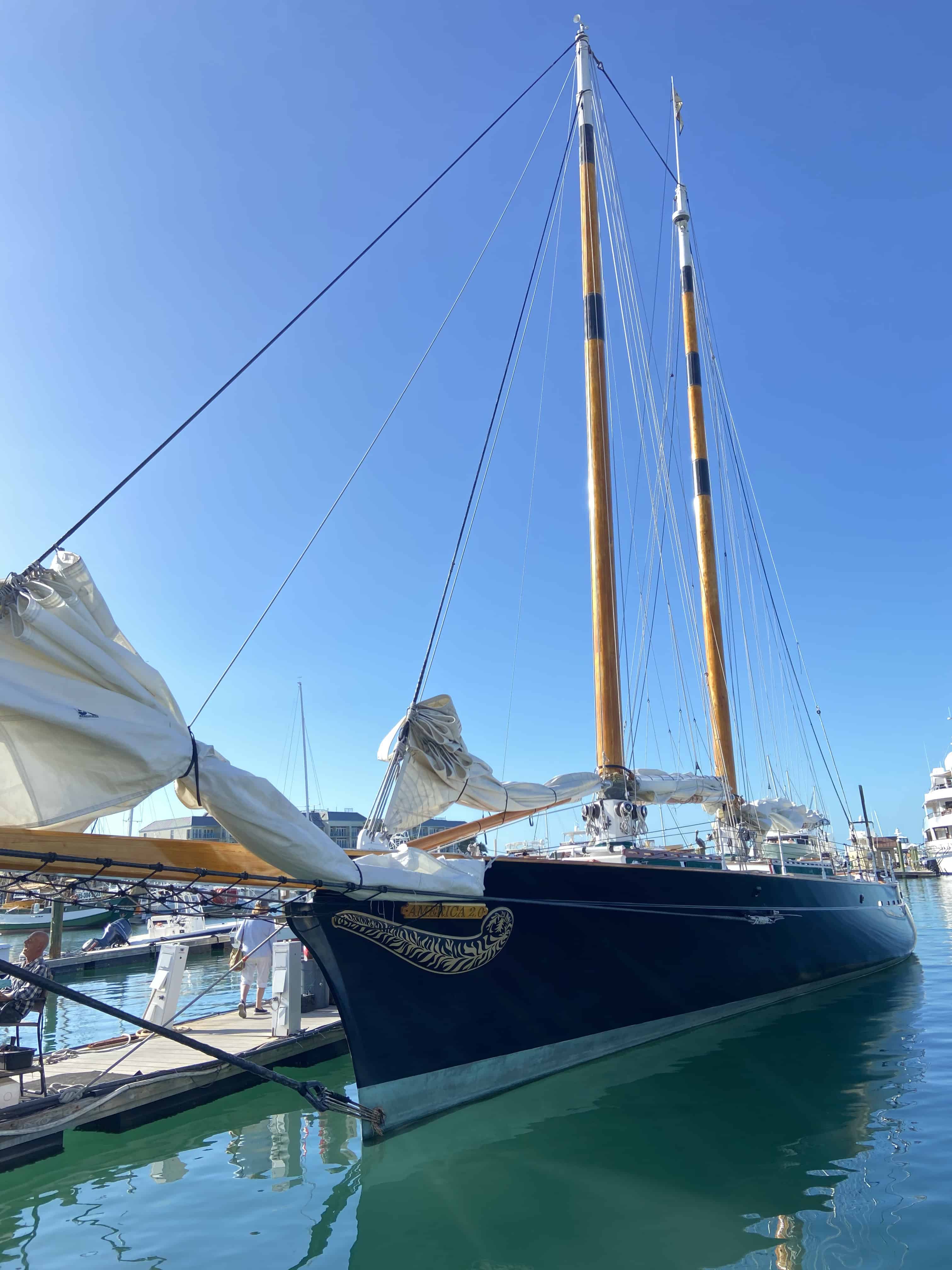 A large, dark blue sailboat with two tall masts is docked in a marina on a clear, sunny day. The boat is the main subject, with calm water and other boats in the background.