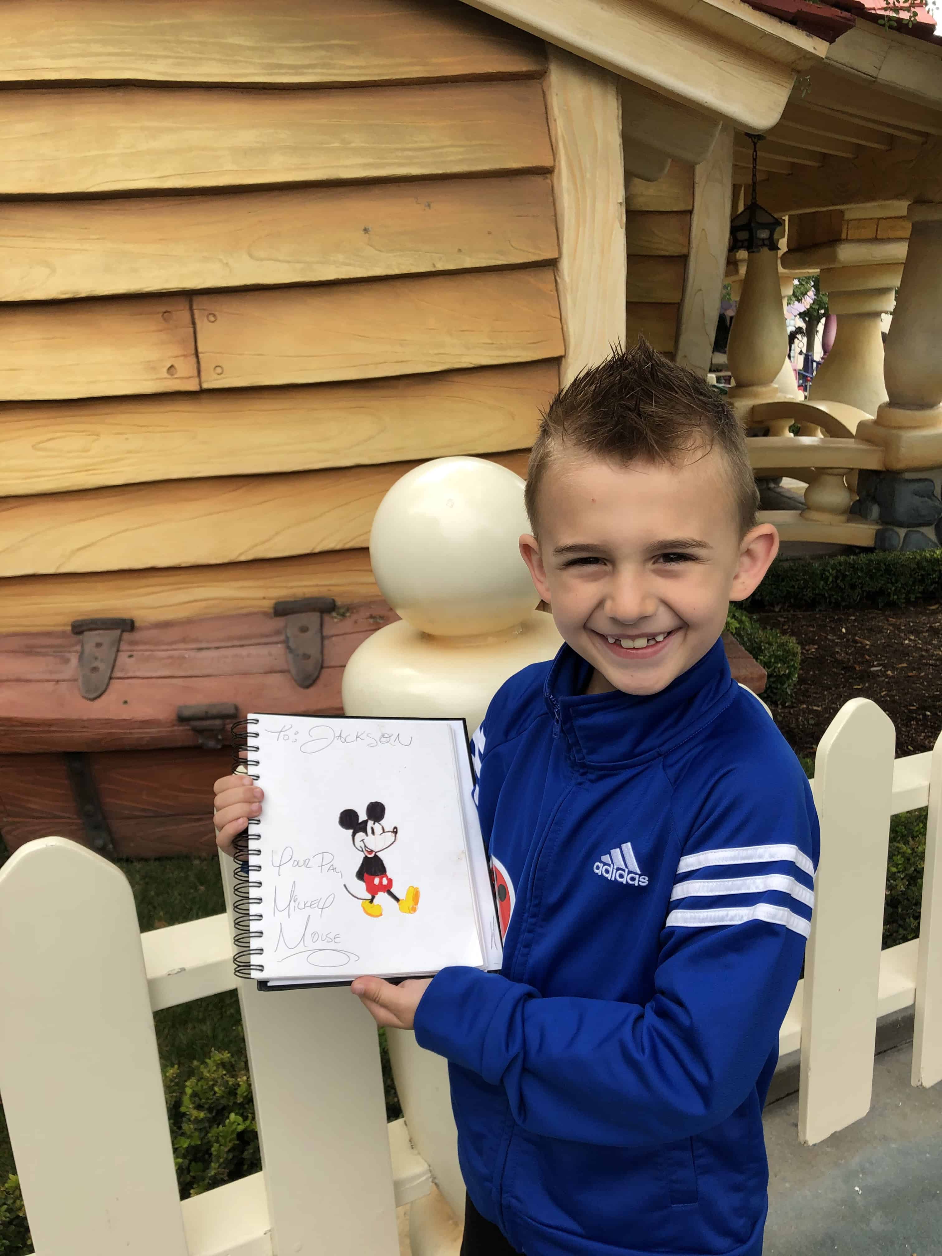 A child holding a drawing beside a wooden building.
