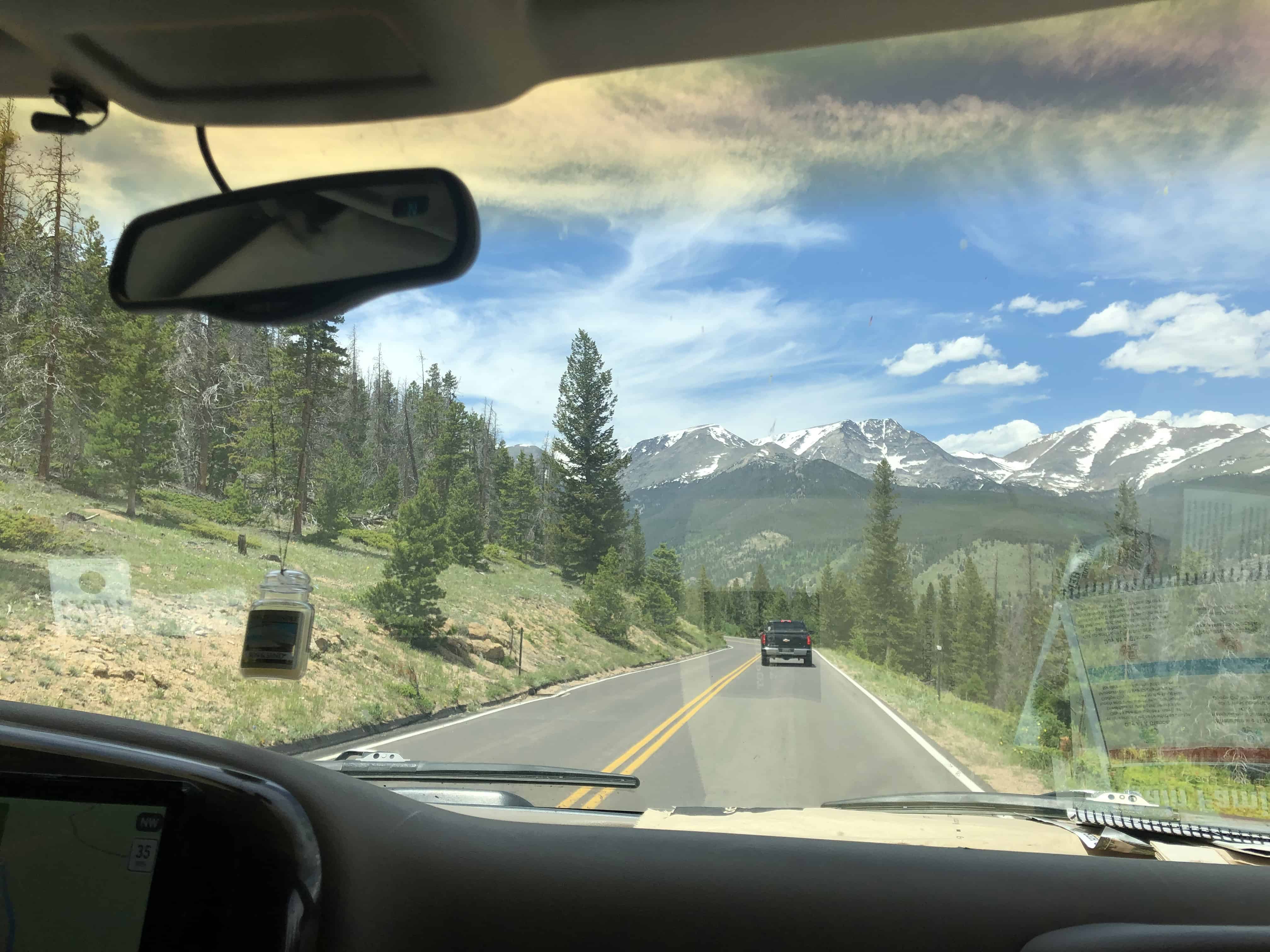 Car view of winding road through pine forest with snow-capped mountains, clear sky, and a rearview mirror.