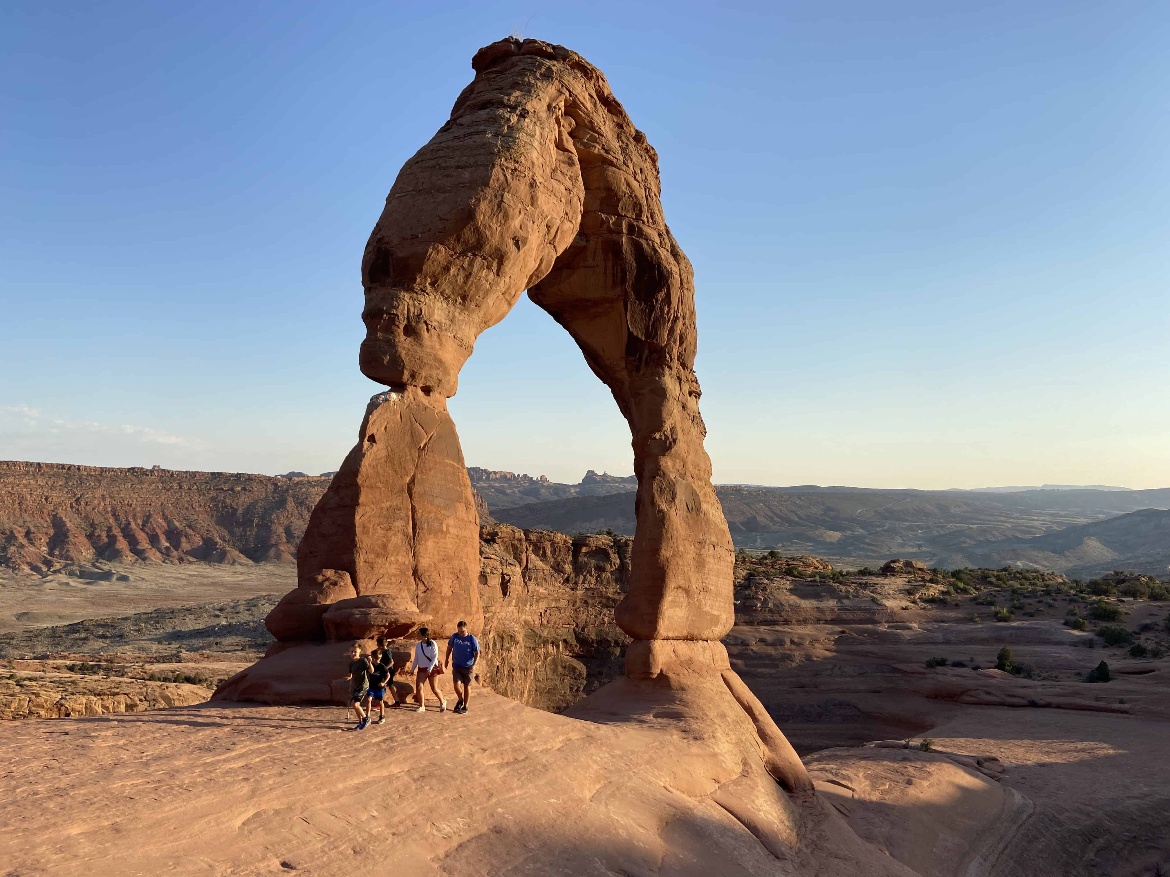 A group of people stands on a rock formation, with a large arch-shaped rock formation in the background under a clear sky.