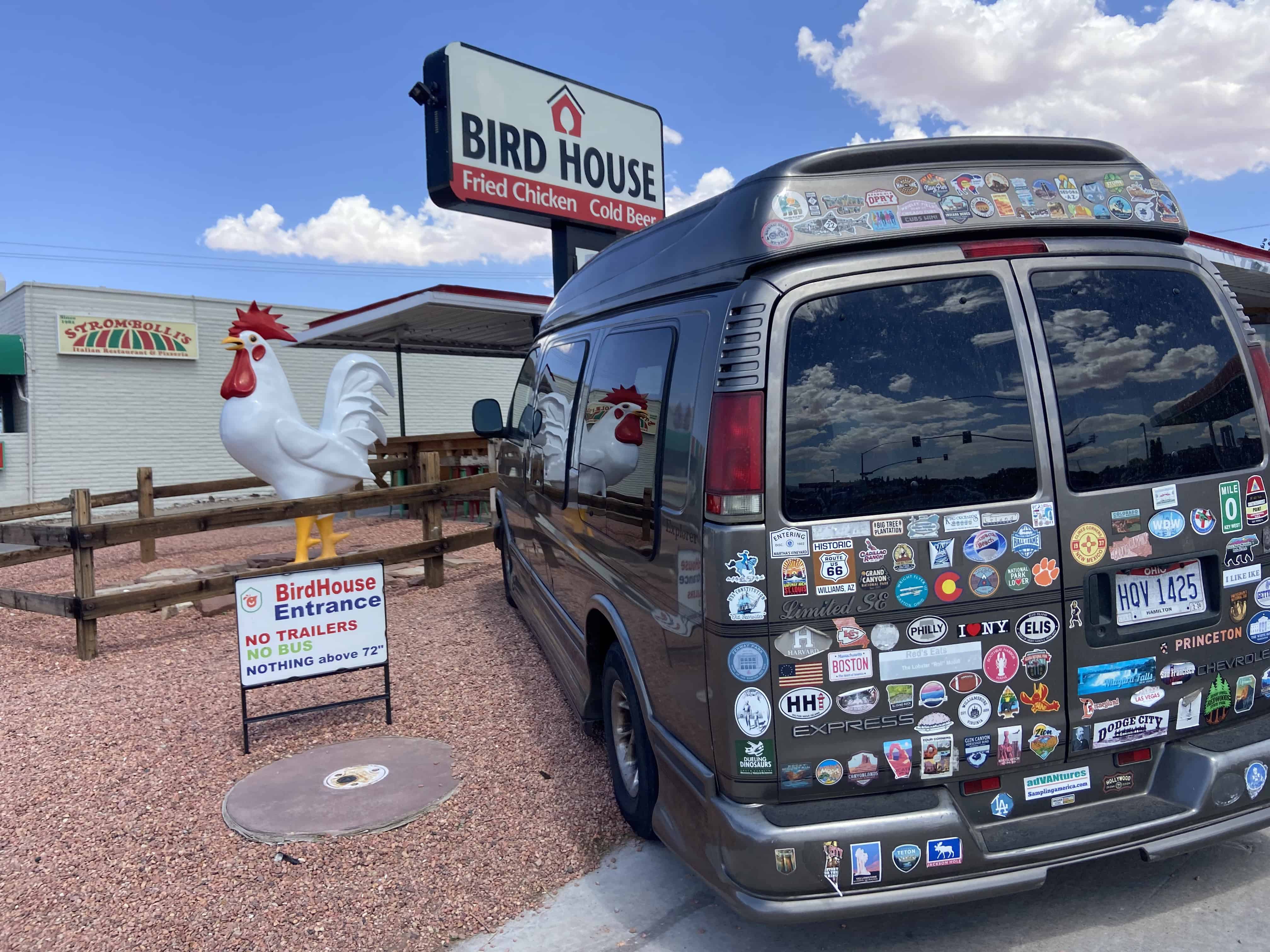 A van with stickers parked at Bird House restaurant, featuring a rooster statue and a sign indicating no trailer access.