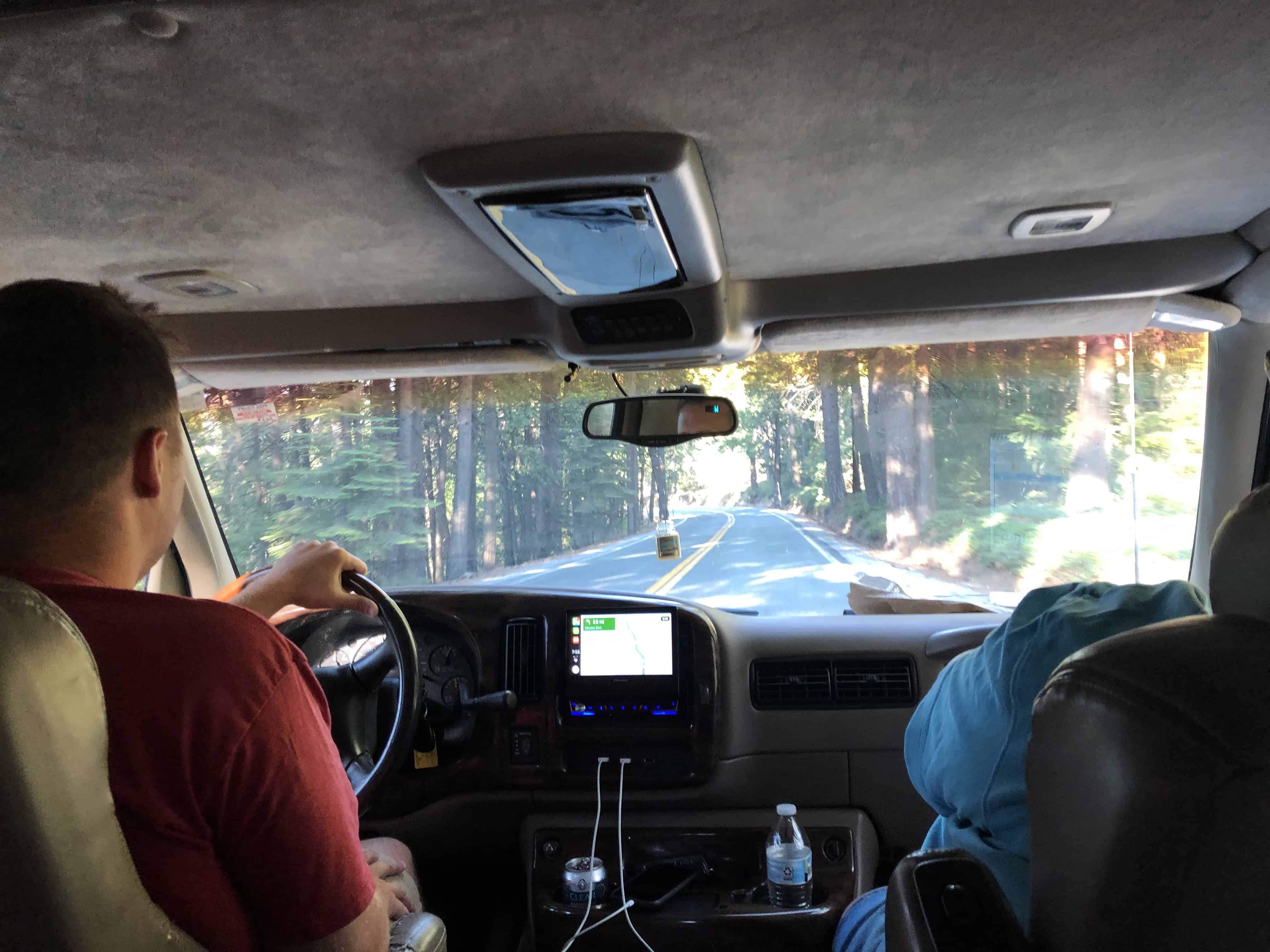 Driver's view of road through forest, car interior with dashboard, seat, and water bottle visible.
