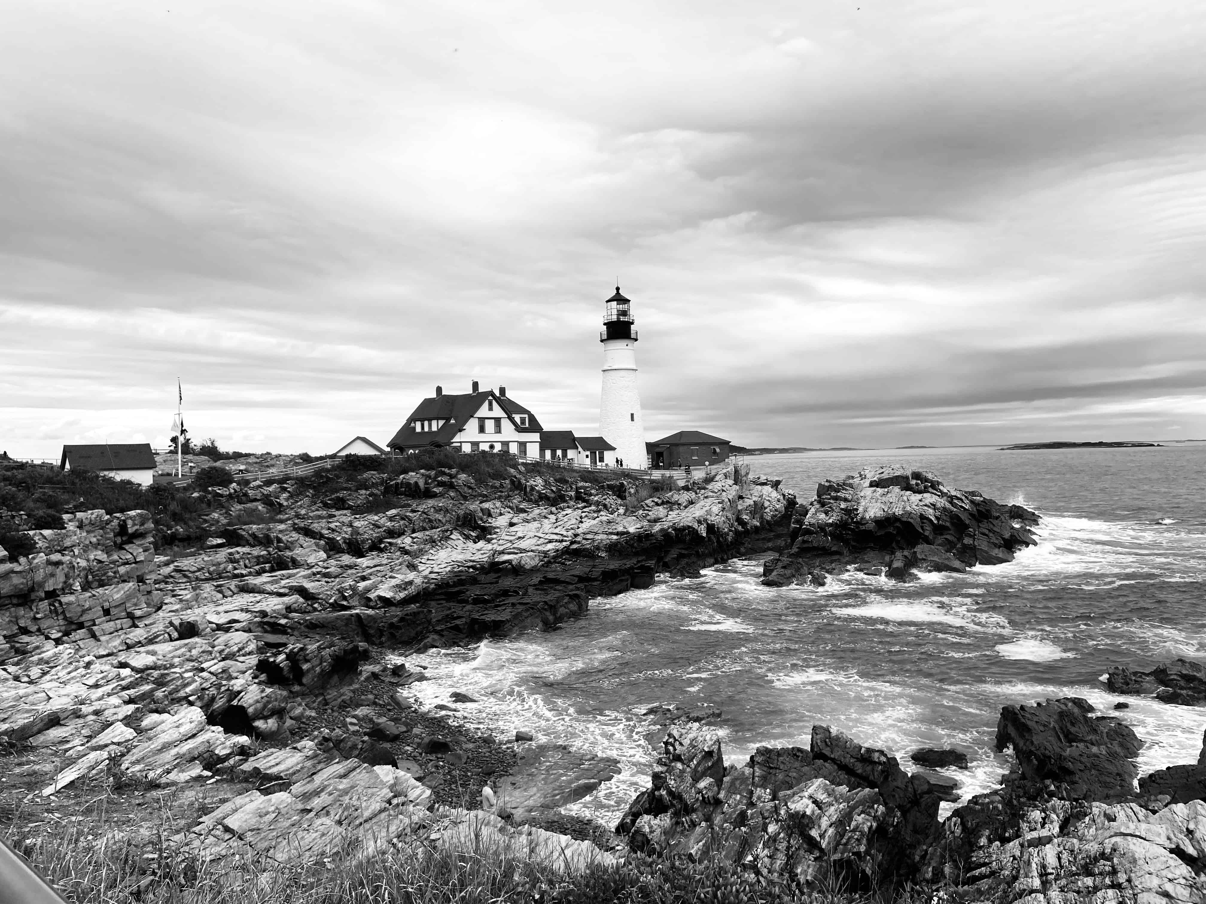 Black and white coastal lighthouse on rocky shore with waves, overcast sky.