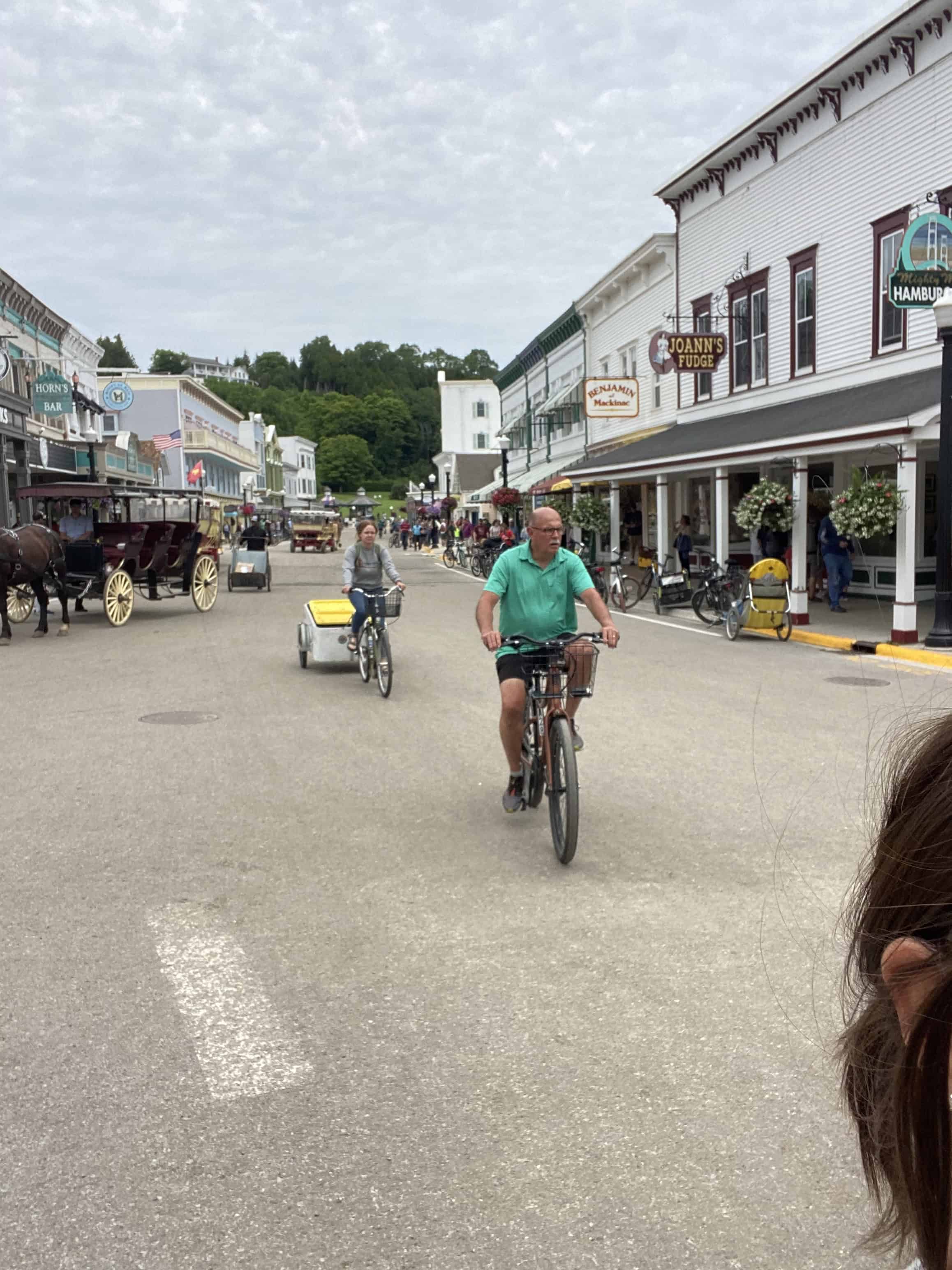 A man rides a bicycle down a town street with horse-drawn carriages and storefronts in the background.