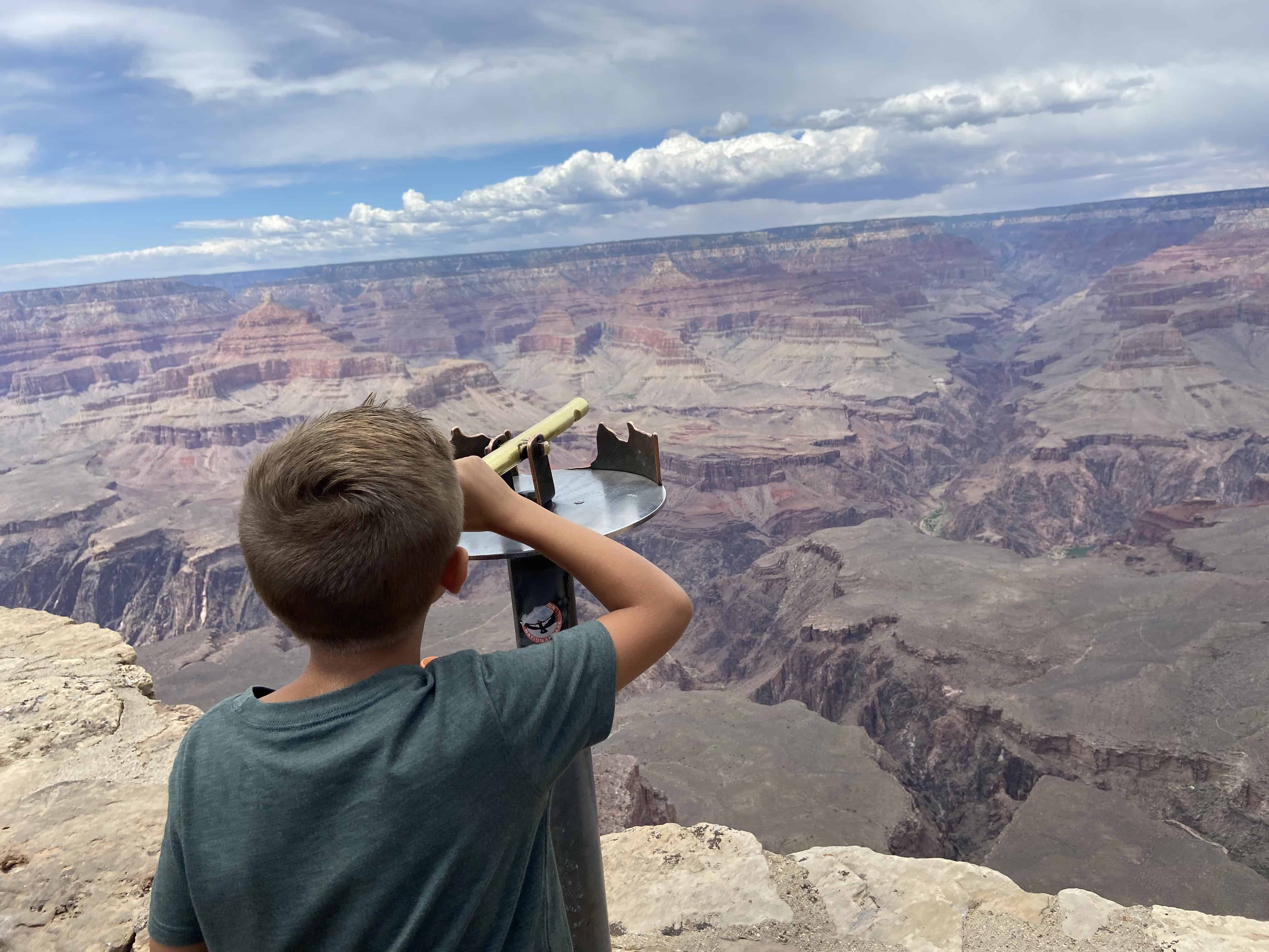 A boy stands on a cliff overlooking a vast, layered canyon, using a telescope to view the expansive landscape.