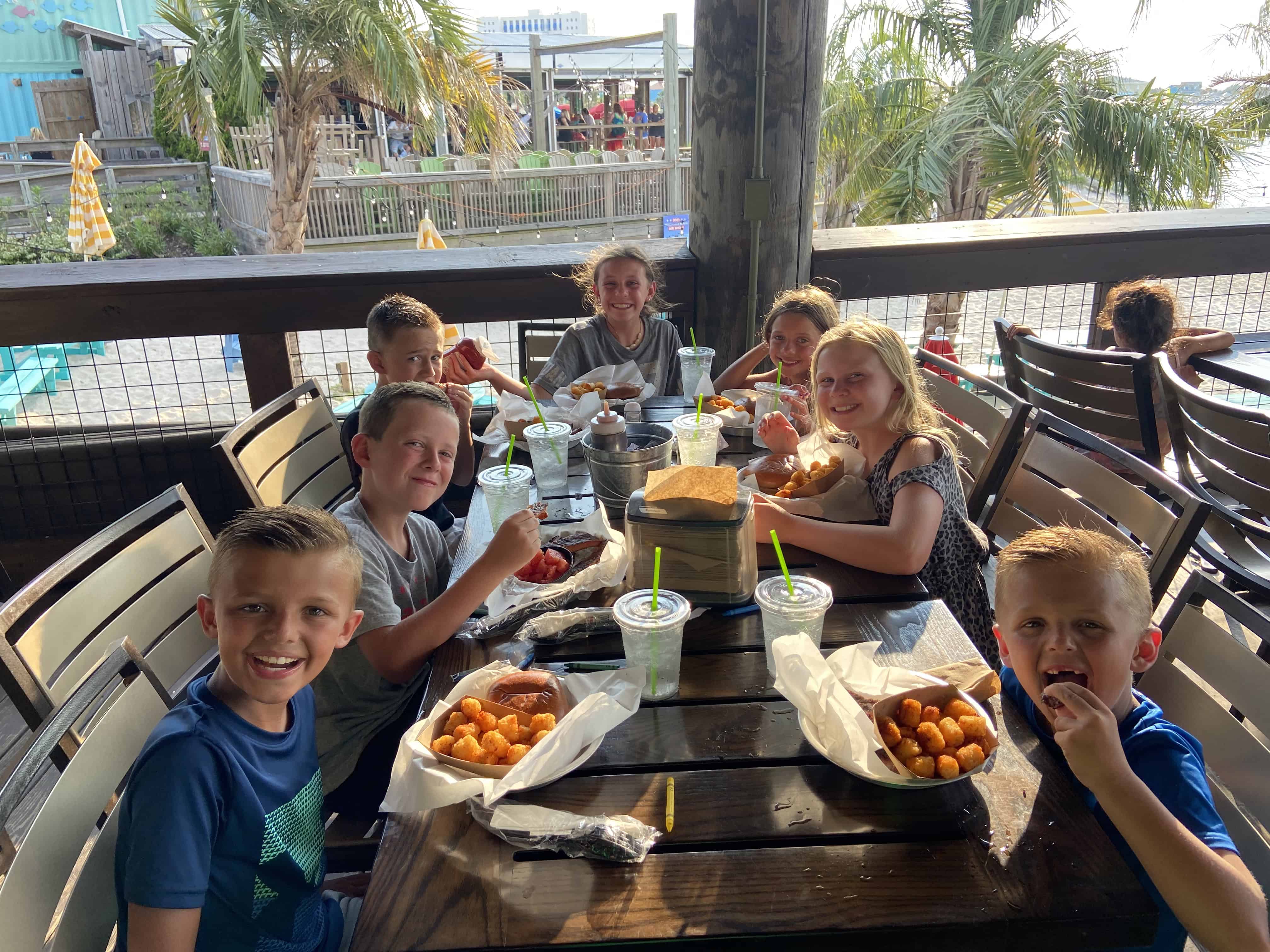 People enjoy outdoor dining at a coastal restaurant, with palm trees and a pool in the background.