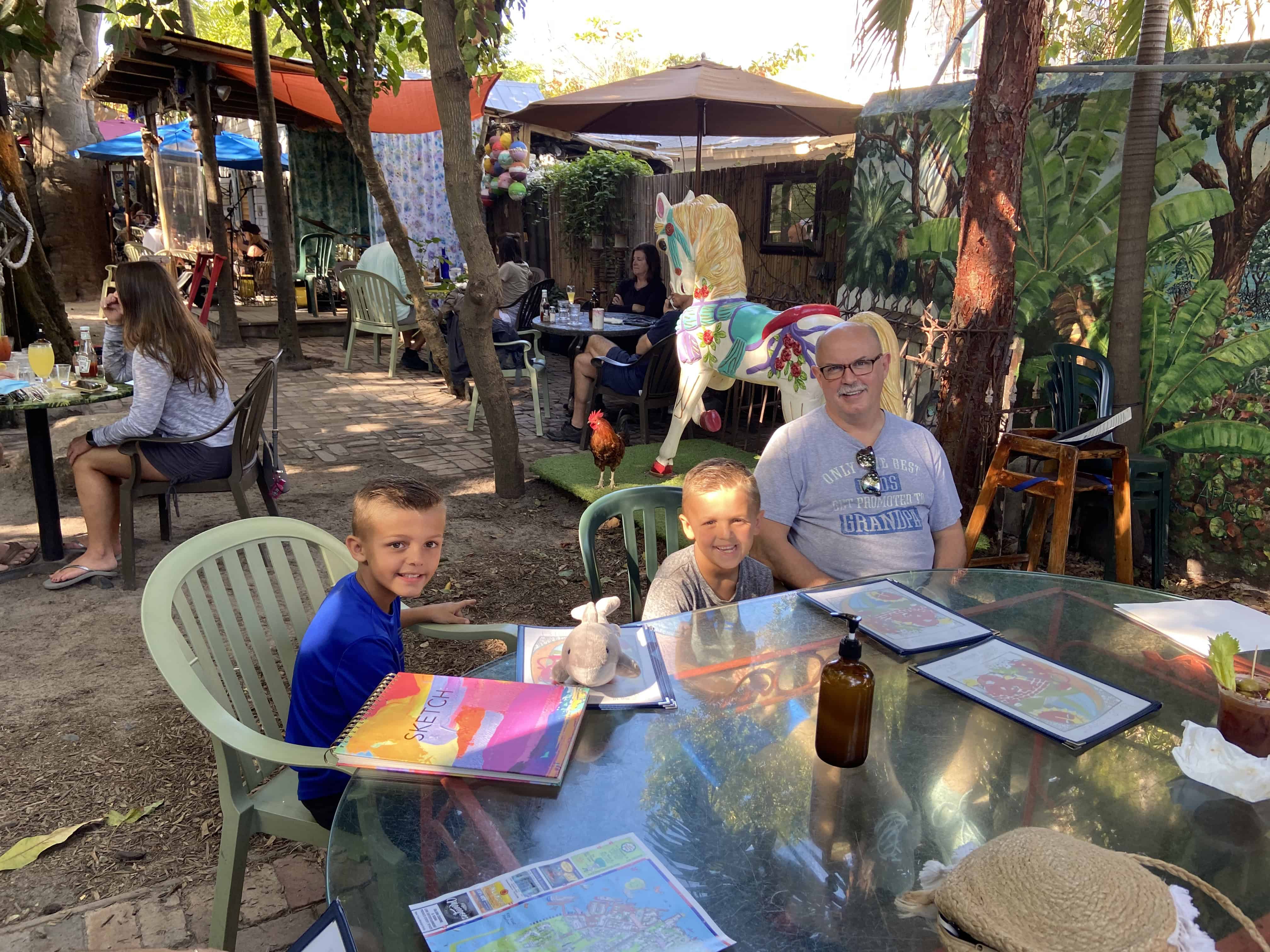 People at an outdoor cafe, seated at a table with menus and drinks, surrounded by trees and other patrons.